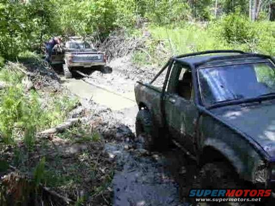 Dark pickup truck with roll bar stuck in mud, another truck being pulled out