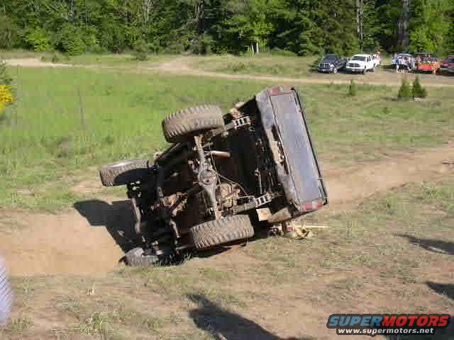 Overturned dark SUV on its side in a dirt pit, with trees and other vehicles in the background