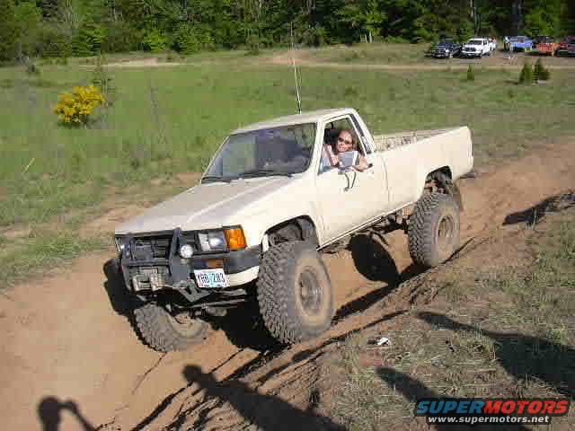 White Toyota pickup truck with large tires driving on a dirt trail. Driver is visible. Other vehicles parked in background