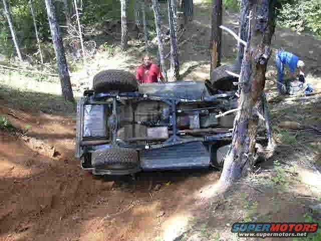 1988 Ford Bronco overturned on its side in a wooded area with two people nearby