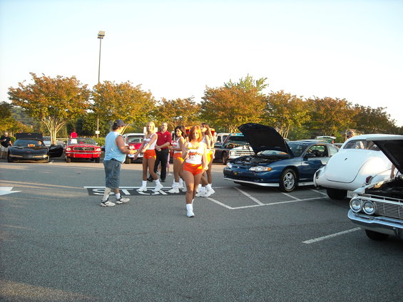 People in orange shorts and white tops stand near cars in a parking lot. One car has its hood open