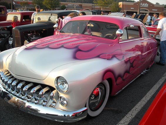 Pink and white custom car with flame graphics, parked at an outdoor event