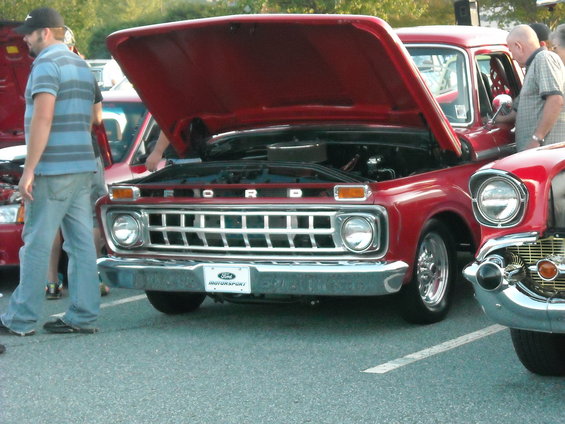 Red 1967 Ford F-100 truck front view, hood open, chrome grille, parked next to another classic car