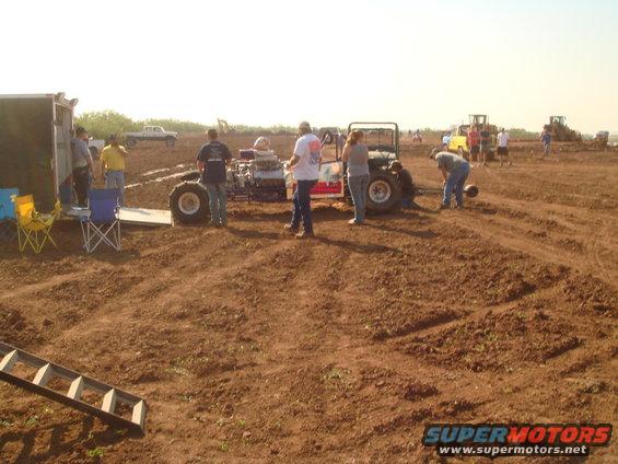 Mud bog racing event with several people gathered around a dragster-like vehicle and a trailer; other vehicles and construction equipment in background