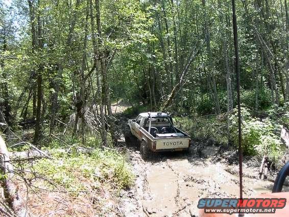 Toyota pickup truck navigating a muddy forest trail. Another vehicle is partially visible in the foreground