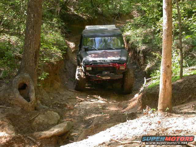 Off-road vehicle navigating a steep, muddy trail in a wooded area. Tires are caked in mud