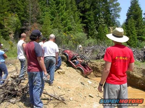 Red off-road buggy stuck in a mud pit, surrounded by trees and onlookers