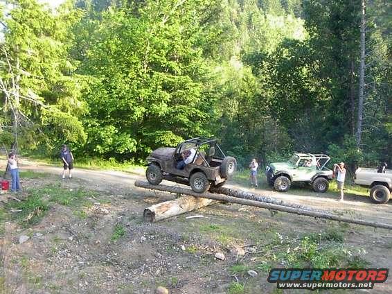 Several off-road vehicles navigate a log obstacle course in a wooded area