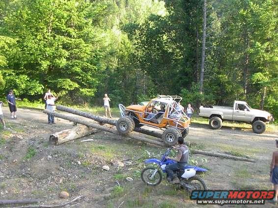 Orange off-road buggy on logs, blue dirt bike in foreground, white pickup truck in background