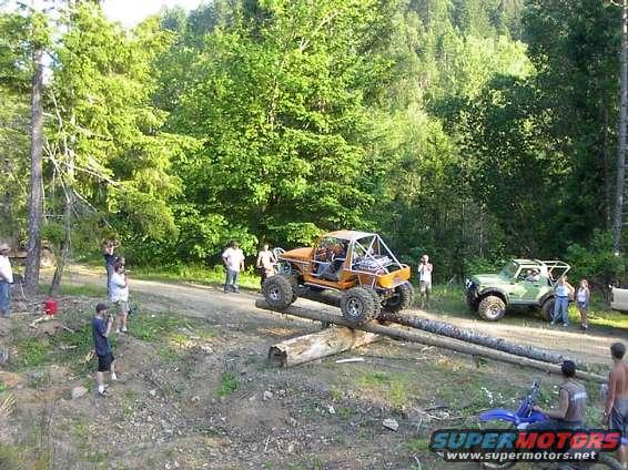 Orange off-road buggy on logs, green off-road vehicle nearby, people watching