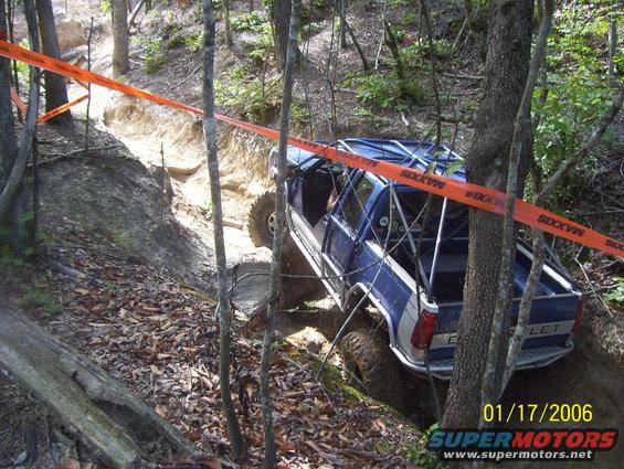 Blue off-road truck with roll cage navigating a steep, wooded trail marked with orange tape
