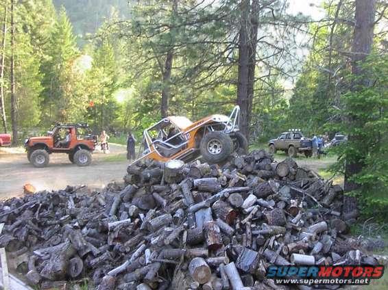 Orange off-road buggy resting on a large pile of cut logs. Another orange off-road vehicle and a pickup truck are visible in the background