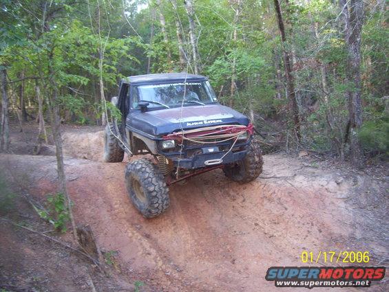 Dark blue off-road vehicle with large tires on a dirt trail, front bumper modified with winch and tow hooks