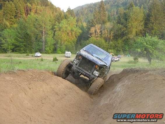 Off-road SUV climbing a steep dirt embankment, tires digging in. Other vehicles and tents visible in the background