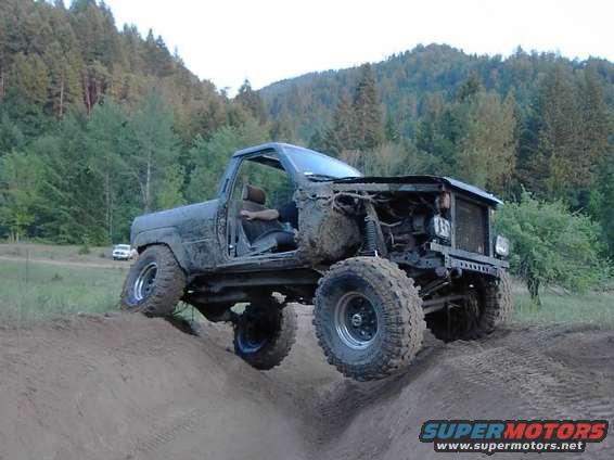 Heavily modified off-road SUV with large tires, lifted suspension, and no front body panels, driving on a dirt hill. A white car is visible in the background