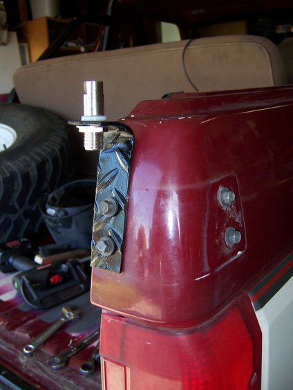 CB antenna mount bolted to the rear corner of a red vehicle, near the taillight. Tools and a spare tire are visible in the background