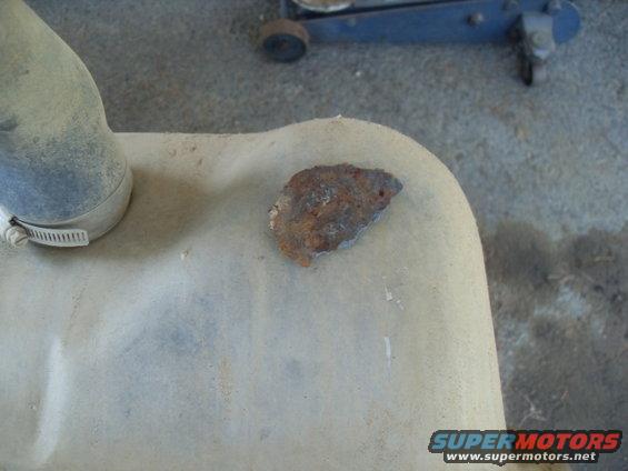 Close-up of a rusty piece of metal on a light-colored surface, possibly a fuel tank, with a hose clamp visible on the left. A blue car jack is in the background