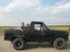 1985 Ford Bronco - Two people ride in a black Ford Bronco on a rural road with fields on either side.