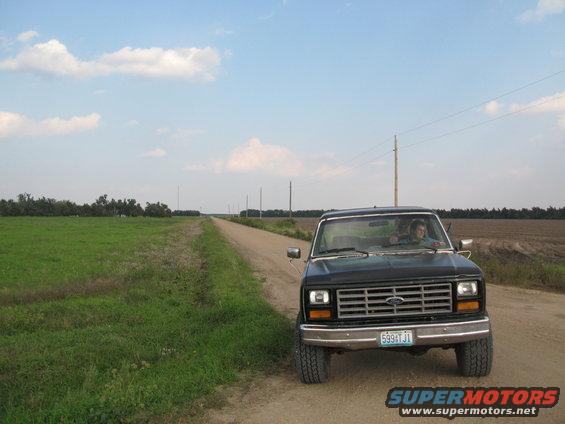Black Ford F-Series truck parked on a dirt road next to a field. Person is driving