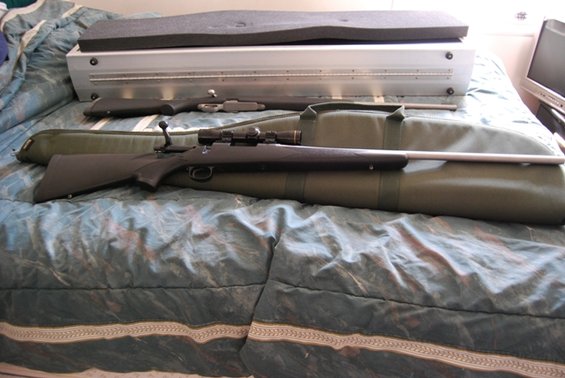 Two rifles, one with a scope, lie on a green gun case on a bed. A metal case with foam padding is behind them