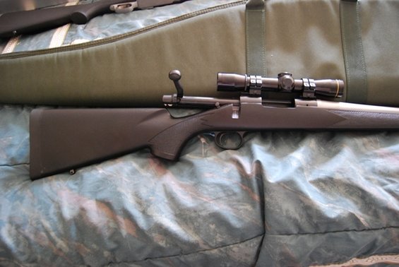 Rifle with scope and bolt action, resting on a patterned blanket next to a green gun bag