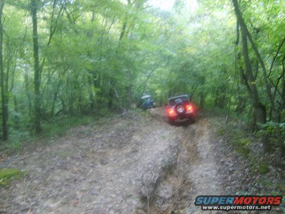 Two off-road vehicles driving on a muddy trail through a wooded area. The rear vehicle has its brake lights on