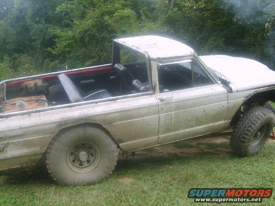 White pickup truck with large off-road tires, missing some body panels, and a roll cage in the bed