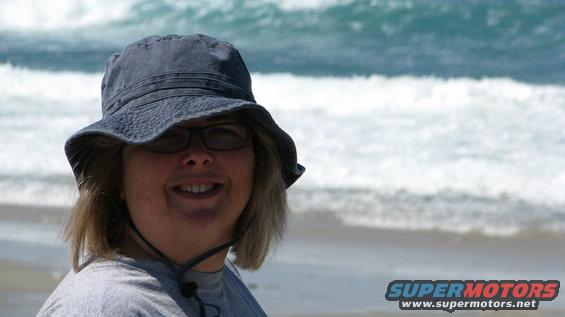 Person wearing a bucket hat and glasses smiles at the beach with waves in the background. Supermotors.net logo visible