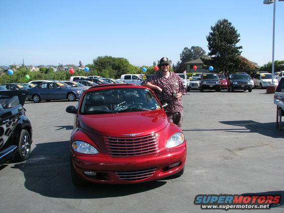 Man leaning on red 2005 Chrysler PT Cruiser at a car lot. Other cars and balloons visible