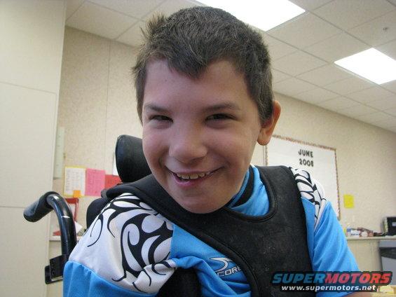 Child in a wheelchair wearing a blue shirt and black vest, smiling