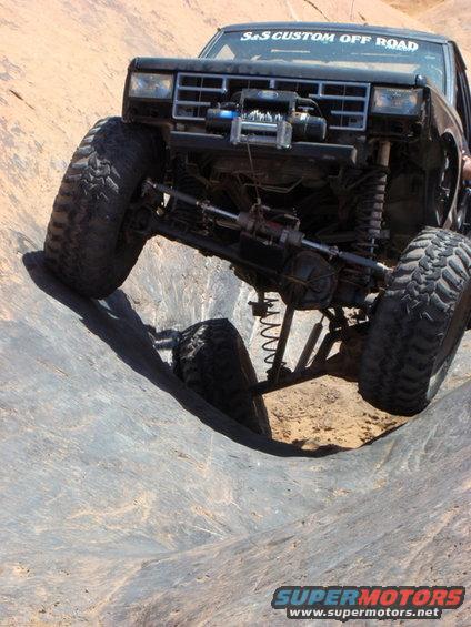 Modified off-road truck with large tires navigating a steep, rocky incline. Winch visible on front
