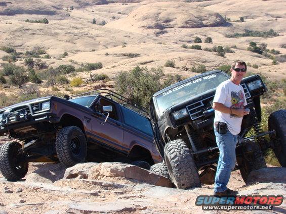 Man standing next to a lifted black off-road truck, with a brown off-road SUV behind it, on a rocky desert landscape