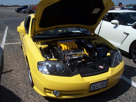 Yellow coupe with hood open, revealing a yellow-topped engine. License plate reads 364-XKG. A white sports car is parked to the right