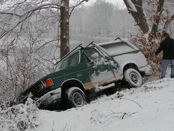 Green and white 1988 Ford Bronco stuck in snow off a bank, with a person standing nearby