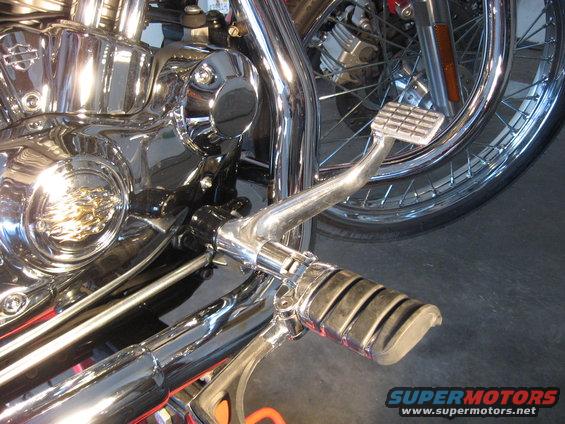 Chrome motorcycle engine and foot controls, with a spoked wheel in the background. The "SUPERMOTORS" logo is visible