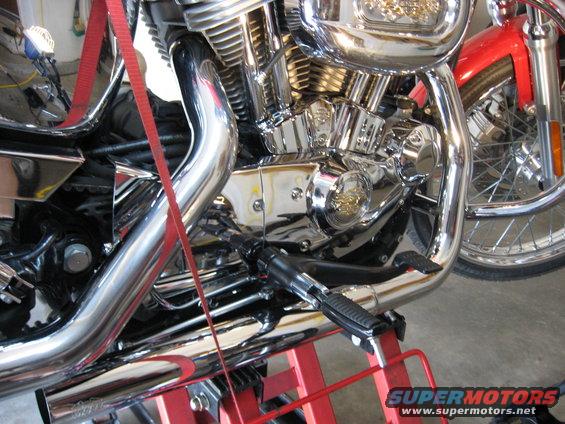 Motorcycle engine with chrome pipes, red strap, and red stand. Visible text: SUPERMOTORS.NET