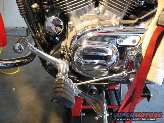 Motorcycle engine detail with chrome covers, shifter, and footpeg. Red strap visible