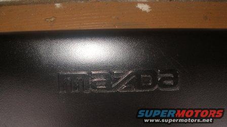Close-up of a black surface with the embossed Mazda logo. Supermotors.net watermark at bottom right
