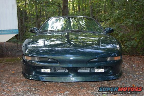 Front view of a dark green coupe with pop-up headlights and fog lights. The car is parked outdoors near trees