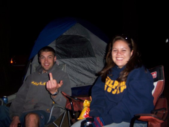 Man gives middle finger while sitting in a camping chair next to a tent; woman smiles beside him