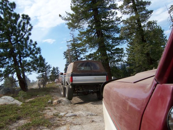 Red truck in foreground, with a 1991 Ford Bronco driving on a dirt road ahead. Trees and sky in background