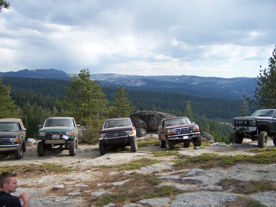 Five off-road trucks parked on a rocky overlook with a forested mountain range in the background