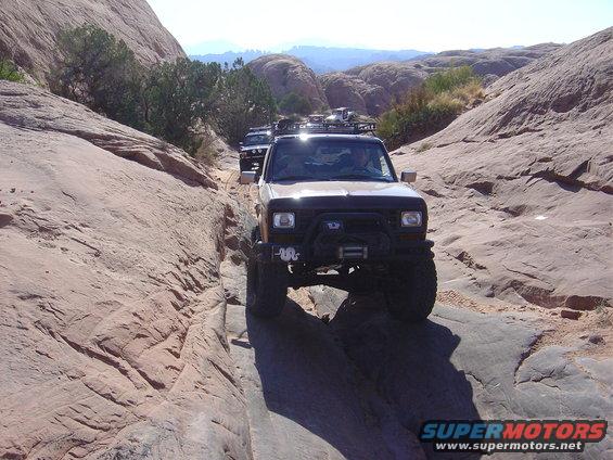 1986 Ford Bronco II and other off-road vehicles navigating a narrow, rocky trail in a desert landscape