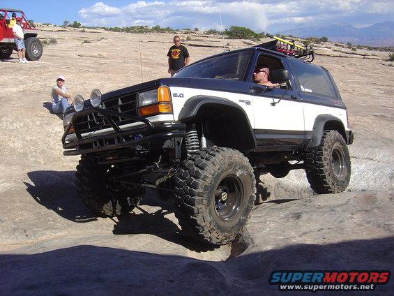 White and blue 1986 Ford Bronco II with off-road tires and a brush guard, driving on rocks