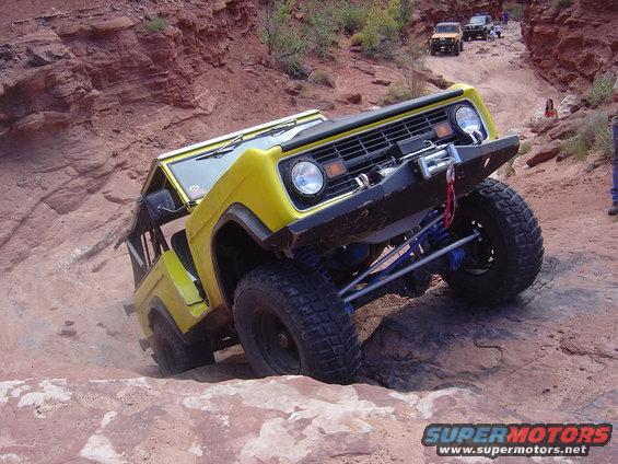 Yellow first-generation Ford Bronco climbing a rocky trail with other vehicles in the background