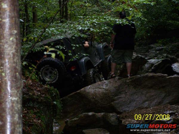 Dark green Jeep Wrangler on rocky terrain, driver's side visible, partially obscured by trees