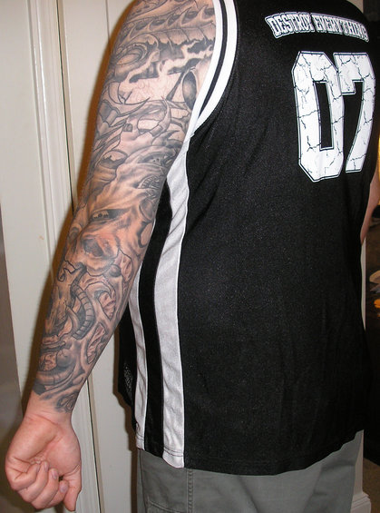 Man's full arm covered in a grey-scale tattoo of mechanical parts and a skull. He wears a black jersey with white stripes and the number 07