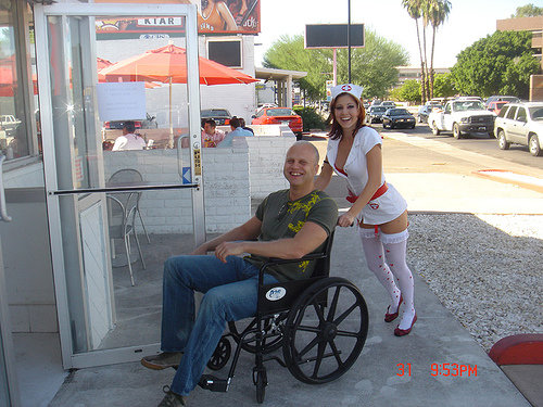 Man in wheelchair pushed by woman in nurse costume, with cars on street in background