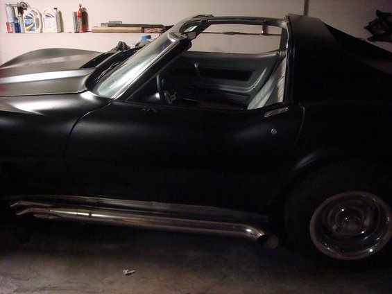 Side view of a black sports car with a matte finish and large chrome side pipes. The t-top roof is removed