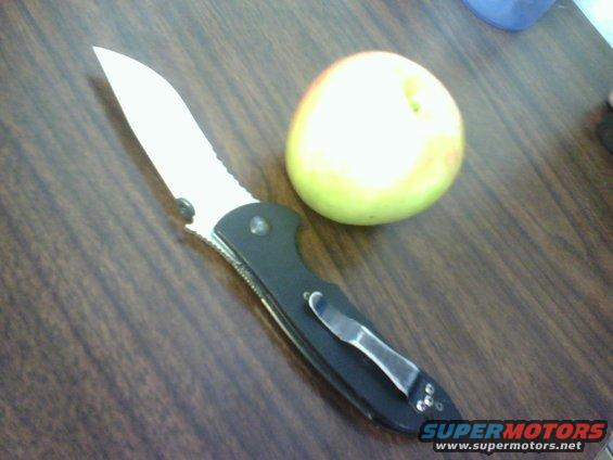 Folding knife with black handle and serrated blade next to a yellow-green apple on a wooden surface